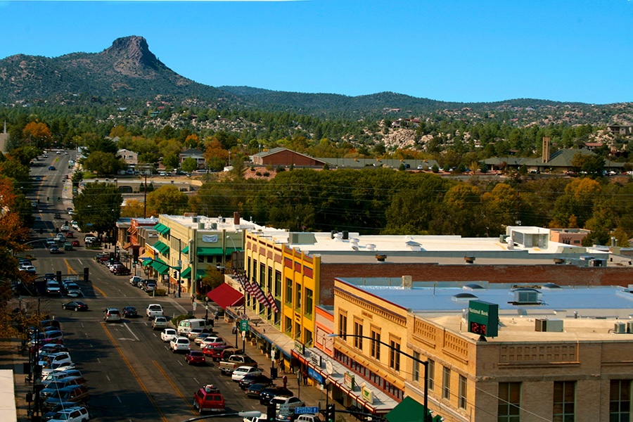 An aerial shot of a main street in Prescott, Arizona, with low-rise buildings featuring quaint historical facades that conjure the authentic small-town charm of the Old West. A pine-covered mountain is visible in the background.