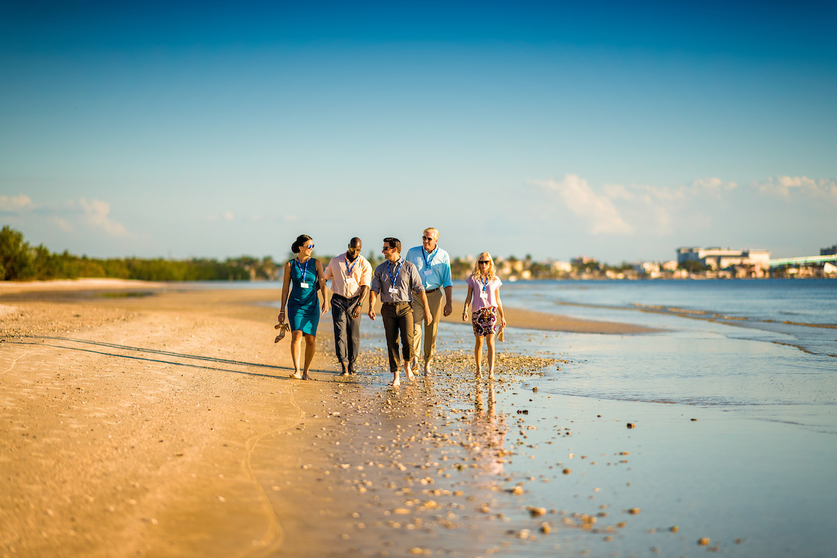 A group of five attendees takes a break from their coastal meeting to stroll down the beach without shoes.