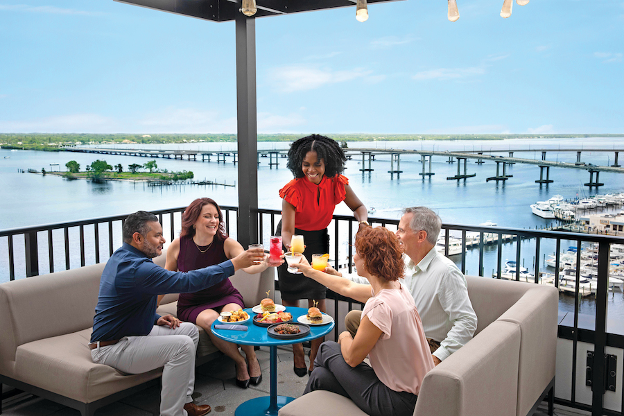 Five meeting attendees sit around a table in lounge seating on a rooftop patio with views of the ocean in the background. They are clinking their drinks in a "cheers." 