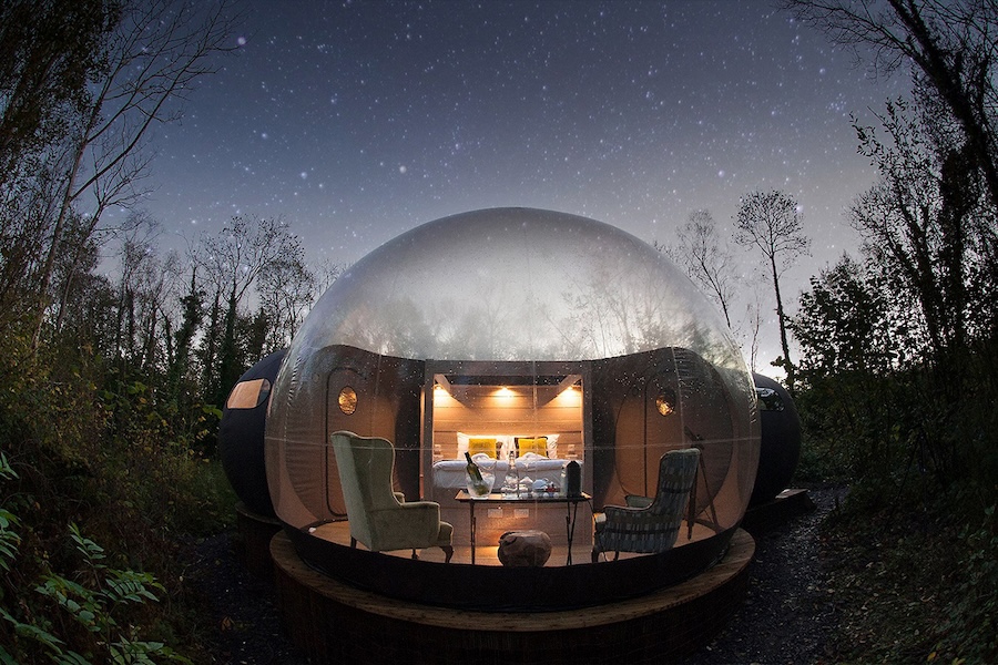 An exterior view of a bubble dome at the Finn Lough Resort in Northern Ireland. A clear spherical structure reveals a cozy living-room scene surrounded by a dark forest at night. The bedroom can be seen through a door leading into a wooden structure at the opposite end of the bubble dome.