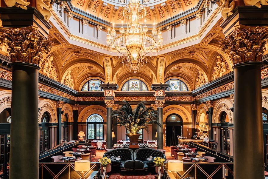 An interior shot of a lavish dining area at the Merchant Hotel in Belfast, Northern Ireland. Neoclassical columns combine with ornate gilded carvings covering the ceiling, while seating consists of plush lounge chairs.
