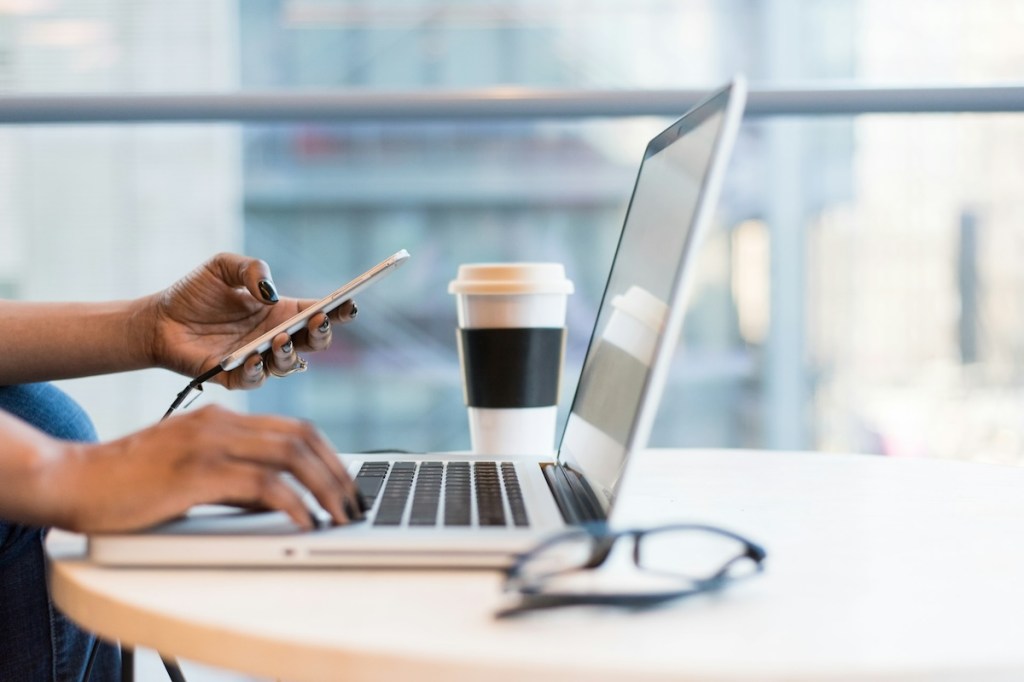 Person using smartphone and laptop with a takeaway coffee cup