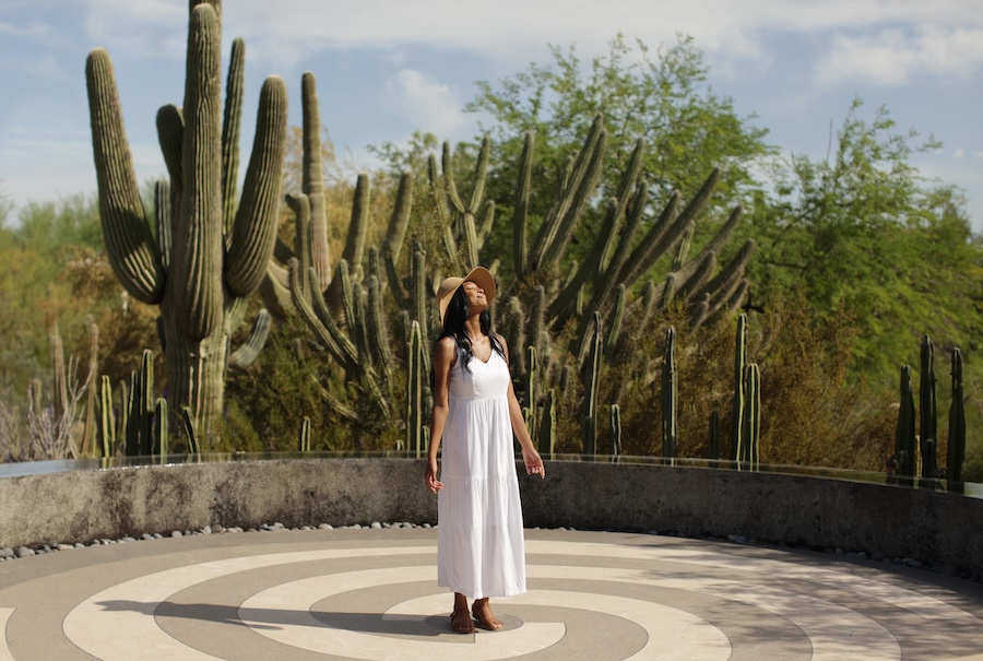 A woman stands in the middle of a circular concrete platform featuring a spiral pattern, with multiple types of cacti and other desert shrubbery in te background.