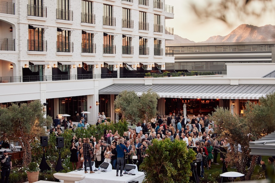 A crowd of people stand in a tree-lined courtyard listening to live music played by two musicians on an elevated stage in the foreground. The clean facade of the new Global Ambassador hotel extends upwards on the left-hand side, while a one-story event space spreads out along the center of the image. A mountain range is visible in the far distance behind the event space.