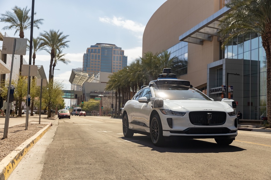 A self-driving car equipped with sensors and exterior cameras cruises along a Phoenix road, with the Phoenix Convention Center visible in the background.