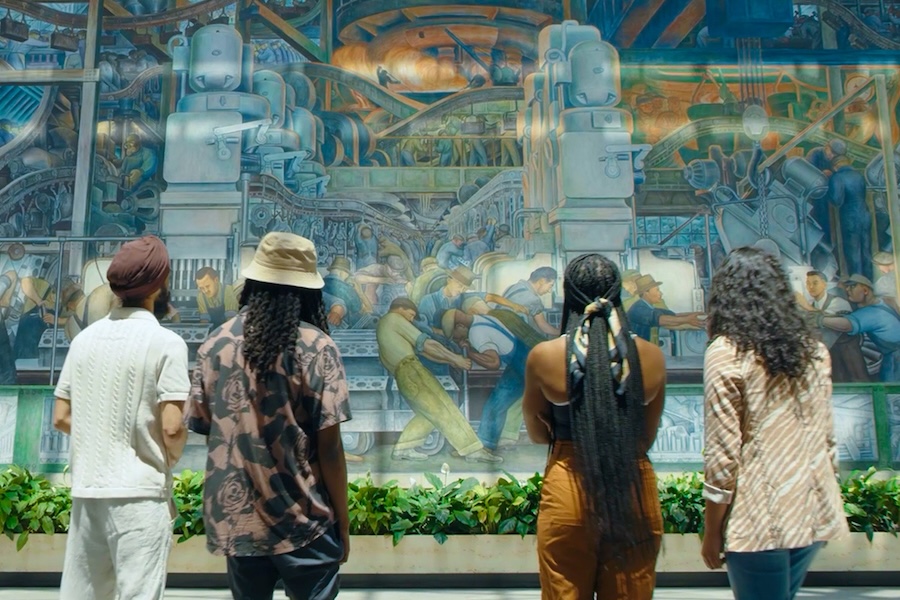 An image capturing the backs of two men and two women who are looking at a mural of industrial workers by Diego Rivera.