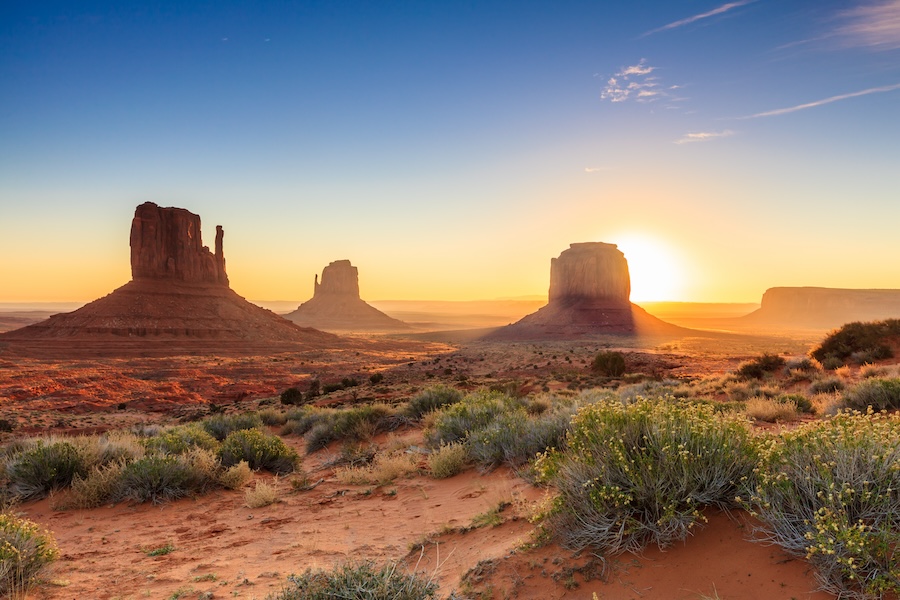 Three rectangular red sandstone buttes line the desert horizon, with a longer plateau mesa in the distance on the far right-hand side.