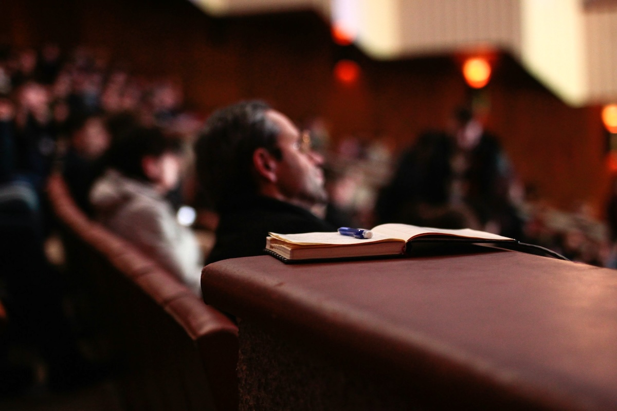 People sitting in lecture theater listening to a speaker with a notebook and pencil in the foreground