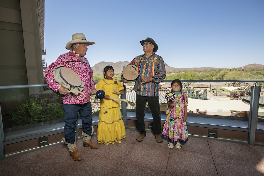 Two men and two little girls stand on the edge of a balcony overlooking the desert landscape at the We-Ko-Pa Casino Resort, a unique venue for meetings. They are wearing a combination of traditional tribal clothing and cowboy attire as they play a mix of percussive instruments — rattles, drums, and sticks.