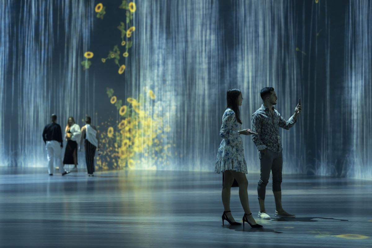 Five people gaze in wonder at projection images of rain and falling flowers on the walls of a large interior space within Superblue Miami. It is part of the Universe of Water Particles Transcending Boundaries exhibit by teamLab.