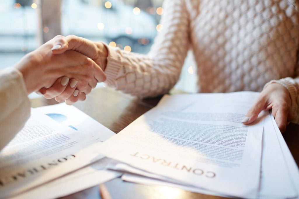 Handshake over desk filled with contracts.