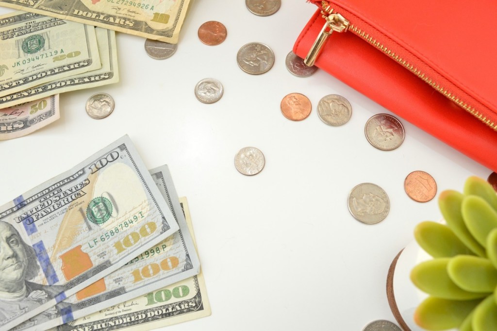 Bills and coins on a table next to a red purse and a plant