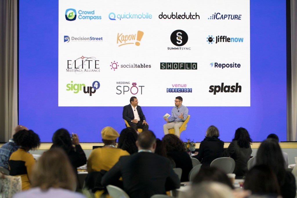 Two men on stage at a conference with a screen behind them showing multiple logos