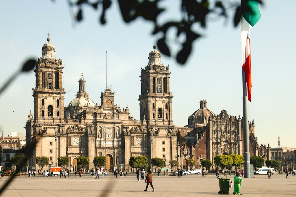 Daytime photo of the Metropolitan Cathedral In Mexico City with a Mexican flag in the foreground.