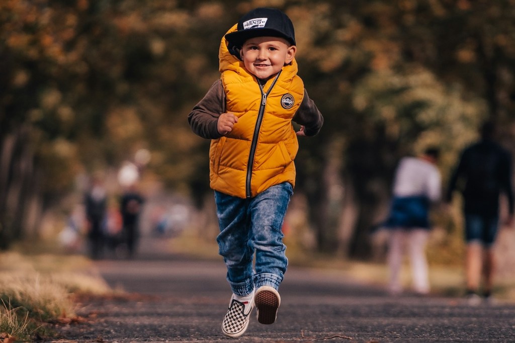 Happy boy walking in park wearing a puffy yellow jacket, cap and and sneakers