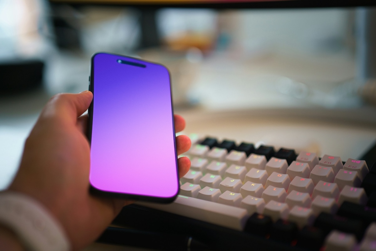 Person holding a smartphone with a blank screen colour gradient in front of a computer keyboard