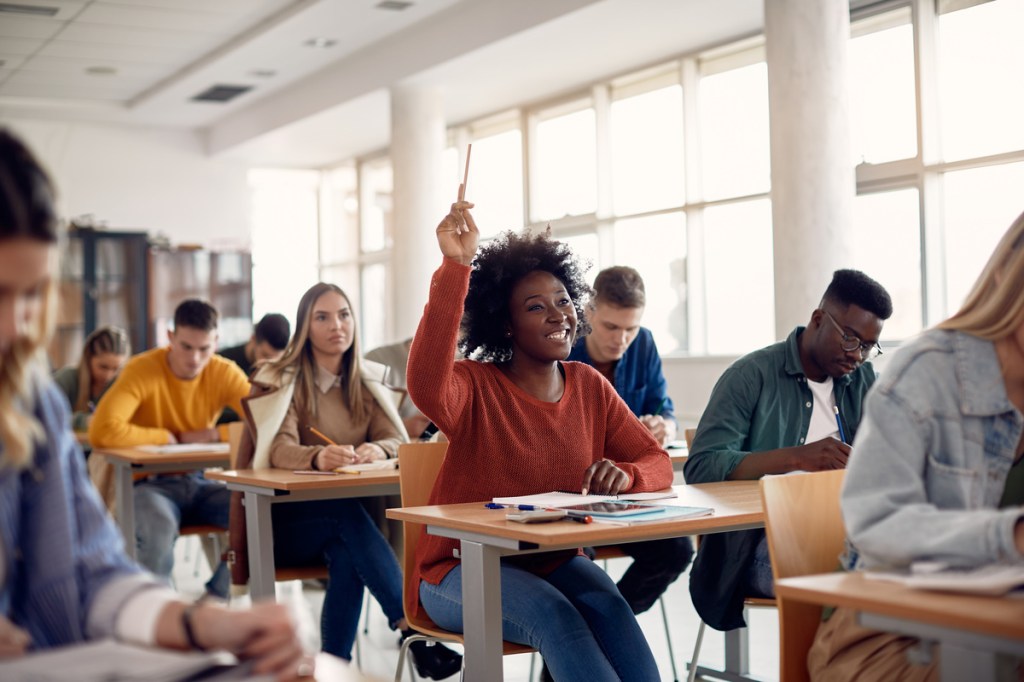 Happy student raising arm to answer question while attending event planning class with her university colleagues.
