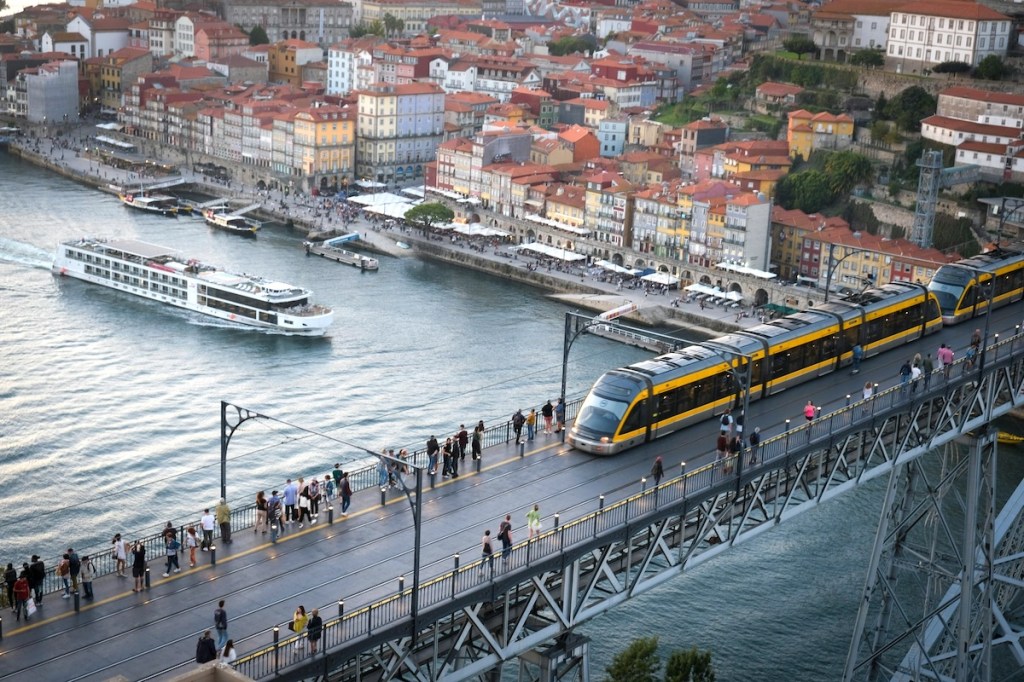 Cityscape of Porto and the Douro River with a river cruise boat and the D. Luis bridge with a modern cable car and pedestrians crossing.